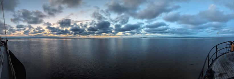 Panoramic photo of a sea almost dry just before sunrise. The cloud-dotted sky goes from yellow on the left to blue on the right.