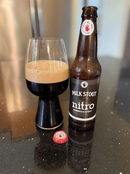A stout glass full of dark beer with a thick tan head is next to a brown bottle with a black label and a bright red left hand on the neck label. The red bottle cap is down front