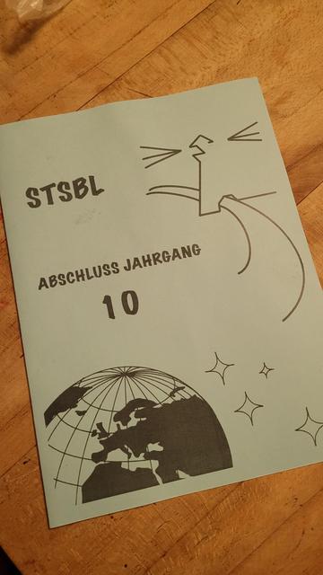 Blick auf eine hellblaue Zeugnismappe auf einem braunem Holztisch. Darauf ein Teil eines Globus und ein stilisierten Leuchturm
 Dazu die Aufschrift:" STSBL (für Stadtteilschule Blankenese) Abschluss Jahrgang 10".