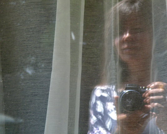 A reflection of myself, a dark haired woman, standing outside looking into my bedroom window holding a camera to photograph the cat and being reflected back