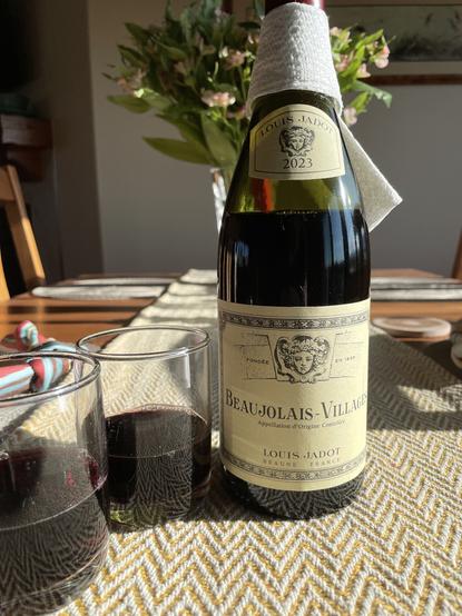 A bottle of Gamay Beaujolais wine with two glasses sitting on a dinner table. There is a vase of flowers in the background and sunshine is coming in from the right side of the picture.