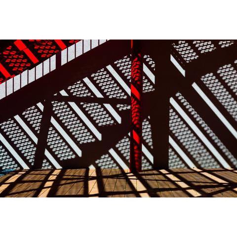 A photograph from under some stairs, painted red. The Sun shining through the mesh steps, creating a high contrast pattern, and dark diagonal lines, on the white cement wall behind it. The ground underneath has a more ochre color.
