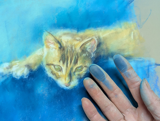 An almost finished soft pastel painting of a brown tabby cat on a blue background along with the painter's hand