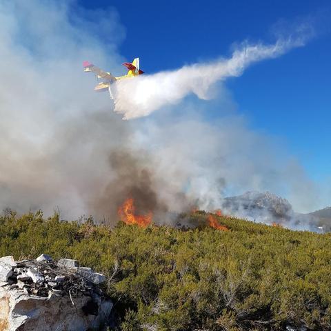A firefighting aircraft releases a large stream of water over a forest wildfire. Thick smoke billows from the burning trees below, with bright orange flames visible among the dense vegetation. In the background, a mountain range rises under a clear blue sky. The landscape is a mix of forested hills and rocky terrain. The scene captures an active effort to control the fire.