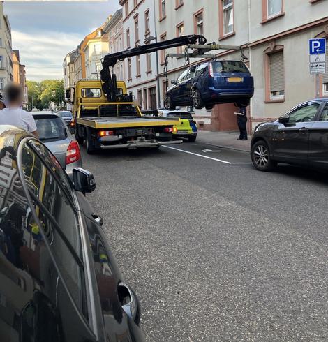 Das Auto mit orangenem Kennzeichen wird per Abschleppwagen vom Behindertenparkplatz gehoben, links im Bild stehen zuschauende Personen