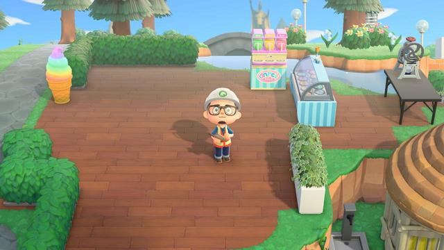 An Animal Crossing: New Horizons character in a construction hard hat and vest stands on a newly laid wooden path plaza. Green bushes and trees are visible in the background, along with a white planter box and a small black table with a machine, indicating a work-in-progress area.
