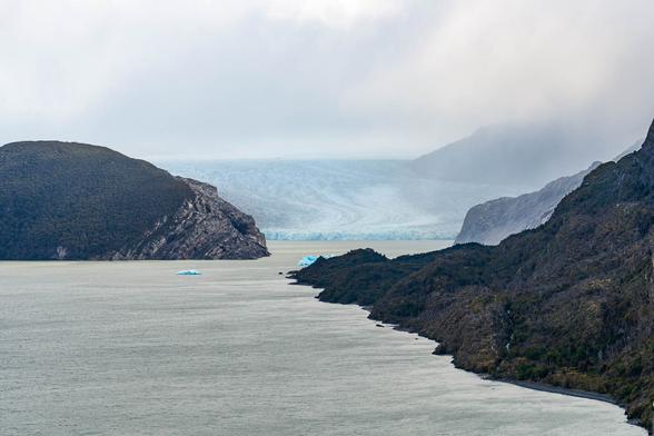 Panoramic view of Glacier Grey under a dramatic sky, seen from a hiking trail above Lago Grey in Torres del Paine National Park. Jagged mountain ridges and moody clouds emphasize the remote and windswept Patagonian landscape.