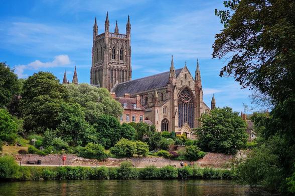 A wide shot of Worcester Cathedral from across the River Severn. The grand, grey stone cathedral dominates the centre, with its tall, ornate tower and large stained-glass window. Lush green trees and foliage frame the cathedral on both sides and in front, obscuring the lower part of its base. A path with a red post box and a bench runs along the riverbank in the midground, with the reflective river water in the foreground. The sky is bright blue with scattered white clouds.