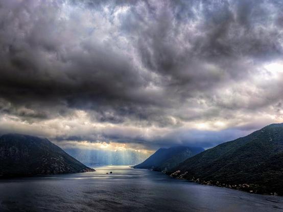 A powerful, high-angle photograph of a thunderstorm forming over the Bay of Kotor, Montenegro. The deep blue-grey water is surrounded by steep, rugged mountains covered in rich green foliage. In the distance, a curtain of heavy rain falls from a thick bank of dark, menacing clouds. Brilliant shafts of sunlight, or crepuscular rays, pierce through the storm clouds, illuminating the downpour and creating a dramatic, ethereal atmosphere over the water. The contrast between the dark storm and the bright sunlit rain is the central focus, capturing a moment of intense and moody weather.