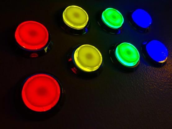 Four pairs of round arcade game buttons, red, yellow, green & blue, lit up with a black background surface.
