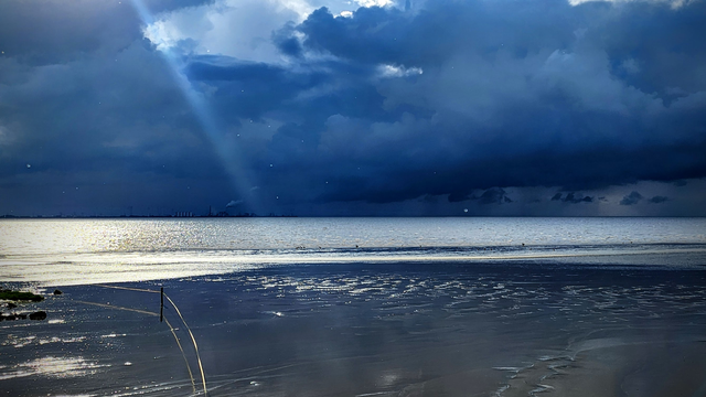 Dunkle Gewitterwolken ziehen über der Nordsee auf. Das ablaufende Wasser und das Watt glänzen weiß im Sonnenlicht, das an einer Stelle noch durch die schwarzblaue Wolkenfront bricht.