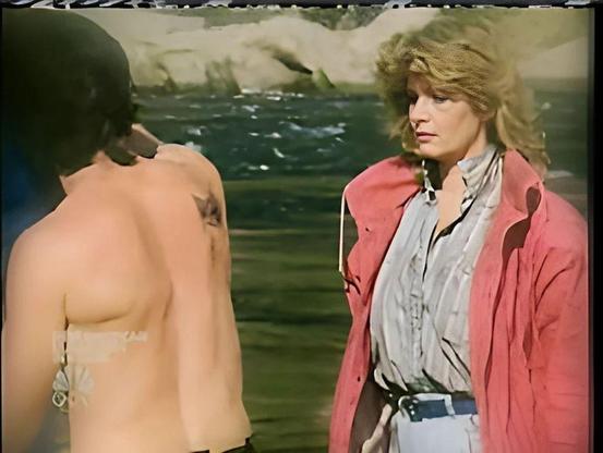 Screenshot from 1986 as Marlena first sees the Phoenix tattoo on John’s shoulder. John stands shirtless, the dark tattoo stark against his left shoulder, while Marlena—dressed in a soft pink jacket and white trousers—looks on with a mix of shock and recognition. They’re outdoors by a rocky shoreline, the moment charged with revelation and history.