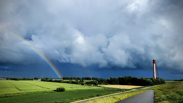 Vom Deich aus geht der Blick ins Hinterland, wo gerade ein Gewitter abzieht. Die Wolkenfront und die Felder, Wiesen und Buschreihen werden von der Abendsonne hell erleuchtet. Ein Regenbogen spannt sich in schillernden Farben auf und am rechten Bildrand ragt ein rotweißer Leuchtturm auf. Das helle
Rot, Weiß, Grün, Beige und die Regenbogenfarben bilden einen harten Kontrast vor den graublauen Wolken.