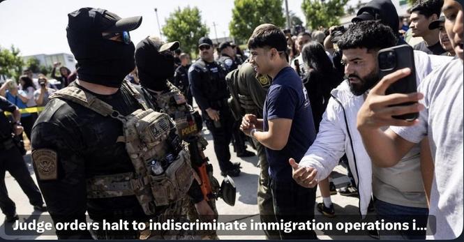 A group of law enforcement officers, some in tactical gear and masks, confronts a crowd of protesters. A few individuals appear to engage directly with the officers, with several people recording the scene on their phones. The atmosphere suggests a tense interaction regarding immigration…