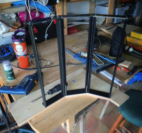 A wooden workshop table cluttered with tools and materials. Lying on its side is a partially assembled Eurorack studio case with black aluminum rails (vertical in this orientation) being screwed onto a precisely cut wooden side panel. Various tools such as a drill, glue gun, and screwdriver are scattered nearby, hinting at ongoing DIY assembly.