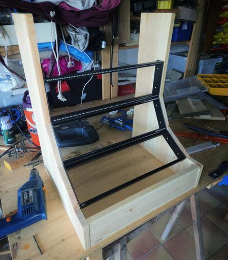 The same Eurorack studio case now standing upright on a wooden workbench. The wooden side panels are fully mounted, and three rows of black aluminum rails are visible, forming a curved structure. The top, bottom, and back panels are still missing, but the intended shape of the 12U, 84HP case is clearly taking form. Workshop tools and materials surround the build in the background.