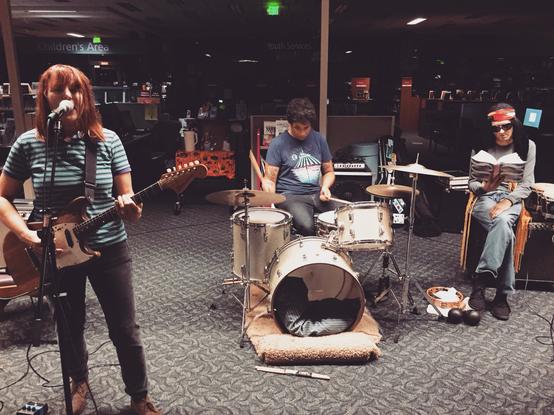 A small band performing in a library setting. The scene features a woman playing electric guitar and singing, a drummer, and a seated individual reading a book. Instruments, including drums and percussion, are visible, and the background includes bookshelves and library decor