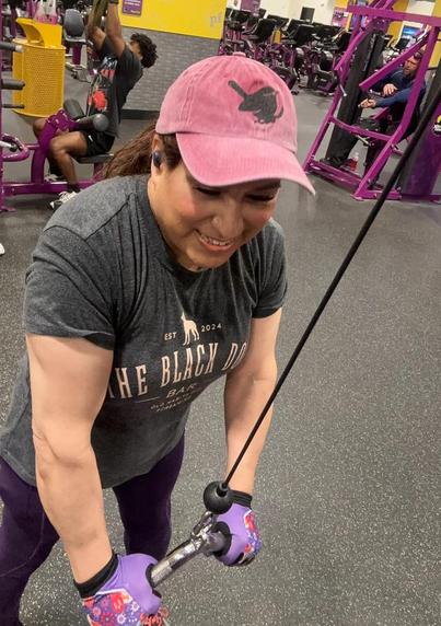 Me, a trans femme Latine woman, at the gym doing a tricep push down.  I'm wearing a pink hat with a black embroidered wren on it, a dark gray t-shirt and purple leggings.  Gym equipment and members can be seen in the background