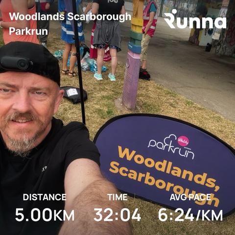 Lee taking a selfie at the "Woodlands, Scarborough" Parkrun sign with other runners waiting for the start of the event in the background.