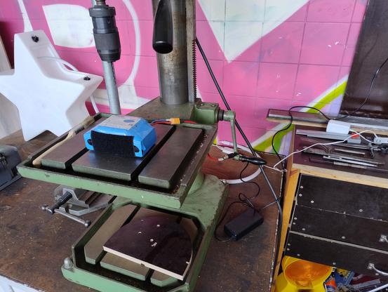 Ein Akkublock der gerade geladen wird während er auf dem Tisch einer Standbohrmaschine mit eingespannten Bohrer liegt.