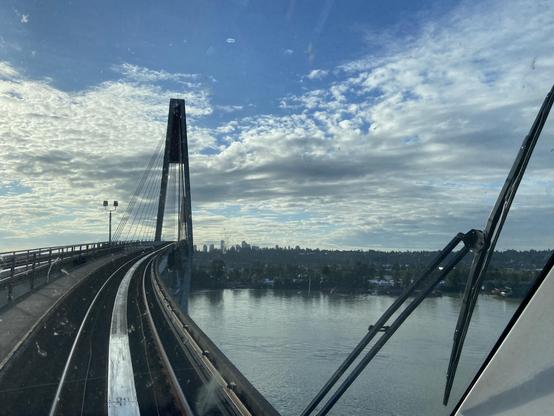 view from the front of a mark three skytrain, approaching the sky bridge. the early morning sun gleams on the dirty windshield and the huge fraser river below ripples from the bridge pillars. skyscrapers in surrey are visible in the distance. partly cloudy sky looks almost as if it were a watercolour painting.