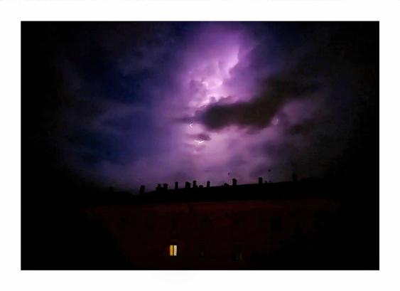 The photo is of a nighttime scene with a dramatic purple-hued thunderstorm.  A lightning strike illuminates the dark, brooding clouds above a silhouetted building's rooftop. There is a single, warmly lit window visible on the dark building a little left of centre in the foreground of the photo.