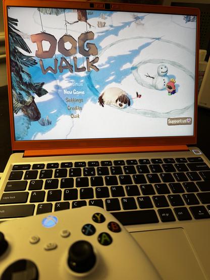 A framework laptop and an Xbox controller in front of a screen with the DOGWALK game loaded. You see a menu to start the game, and a fluffy dog character next to their human and a snowman.