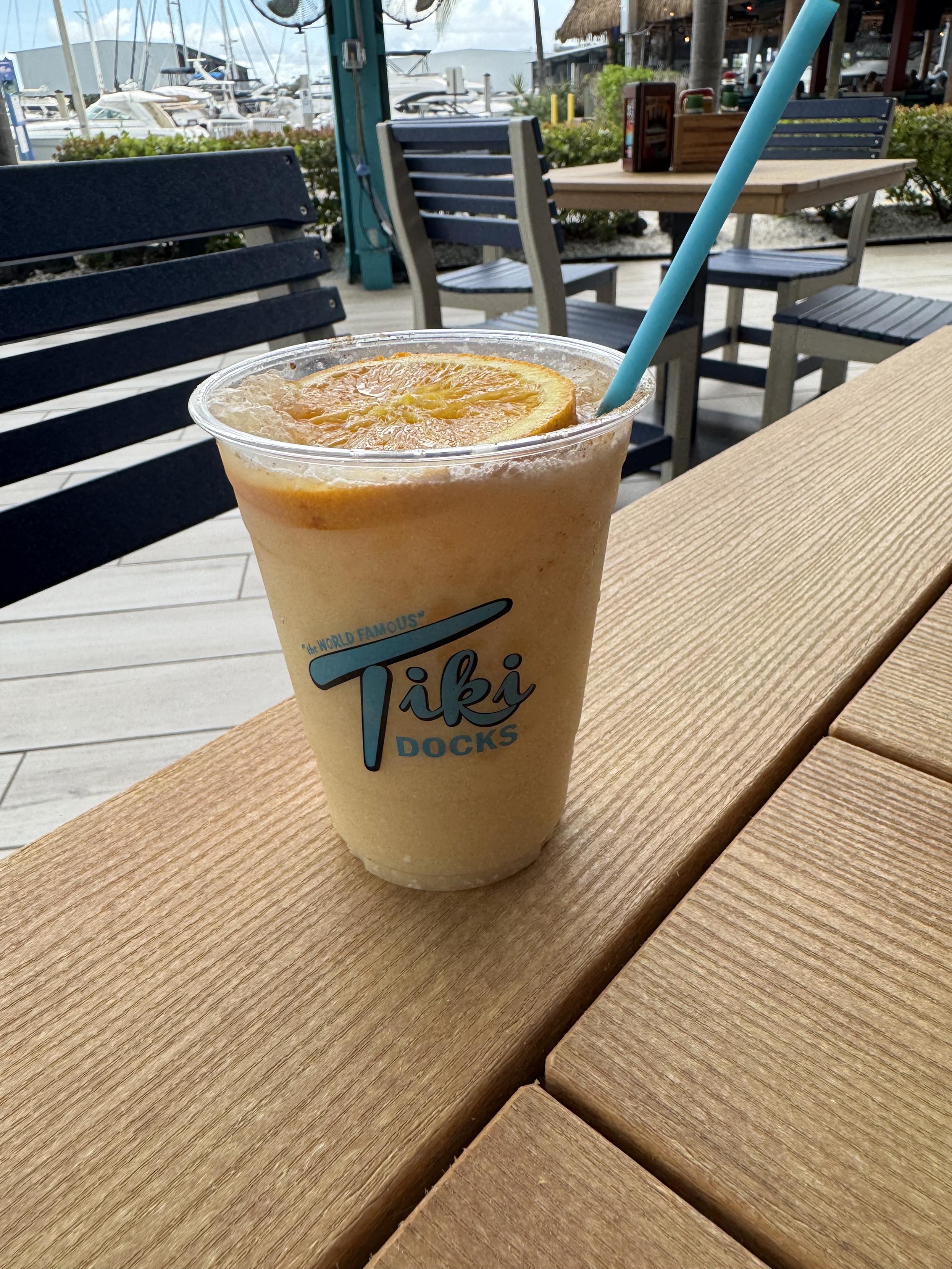 A plastic cup with an orange-cream colored drink in it, garnished with an orange wheel. The cup has a logo reading “‘The World Famous’ Tiki Docks.”