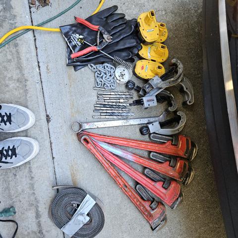 a mess of tools rescued from a dump bin. 4 24" pipe wrenches, many rotohammer bits, some strap, several pipe cutting tools with missing cutters, some dewault batteries, a pile of very heavy duty rubber gloves. my lil grey converse toes peaking.