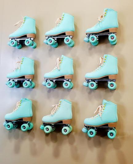 A grid of nine retro-style roller skates is displayed against a solid beige wall. The skates all have the same design: light blue uppers with tan laces and tan accents around the heel. They feature traditional quad wheels in matching light blue, attached to a silver colored frame. The arrangement is a neat 3x3 grid, providing a visually balanced and eye-catching display. The light background provides stark contrast to the vintage design of the skates, further enhancing their presence.