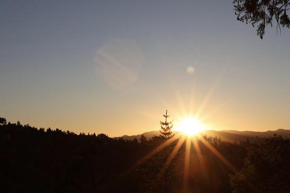 A vibrant sunset is captured behind mountainous silhouettes, with rays of sunlight radiating outward. The sky transitions from blue to warm hues, and evergreen trees are visible in the foreground.