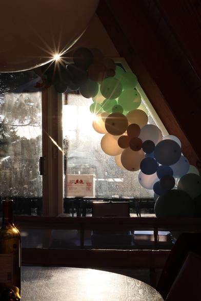 A window with a colorful balloon arch in shades of green, blue, and brown. Sunlight streams through the window and balloons, creating long flares across the balloons. At the upper left is a quarter circle of a large white globe; the sun reflects in a line around the bottom and creates a bright point with 14 bright rays radiating from it.  A bottle of wine is on a table in the lower left foreground, with yet another flare.