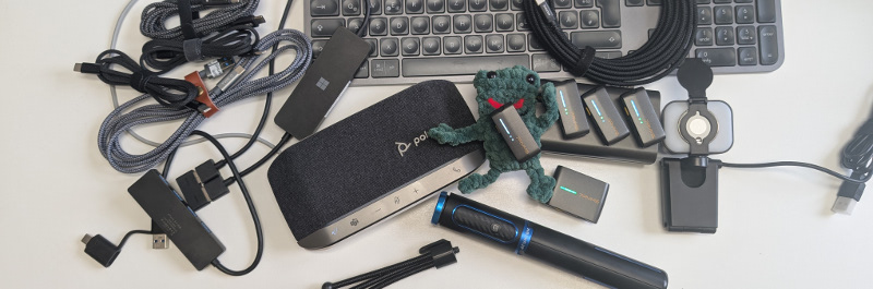 Technische Geräte und Zubehör auf einem Schreibtisch, darunter Kabel, Lautsprecher, Mikrofon, Batterien und Tastatur.