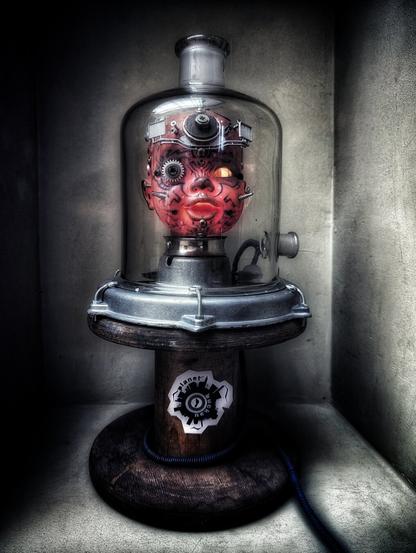 A surreal and eerie close-up of a steampunk-inspired mechanical head encased in a glass bell jar. The head, painted a deep red, features complex clockwork gears and mechanisms on its forehead and around a glowing, sinister-looking yellow eye. A second, smaller eye is visible on the opposite side. The head rests on a metallic base with visible bolts and pipes, which in turn sits on a weathered wooden stand. A sticker with a stylised cogwheel and the word 'Planet Buckau' is affixed to the stand. A coiled blue wire snakes away from the contraption. The background is a dimly lit, textured wall, adding to the unsettling, macabre atmosphere.