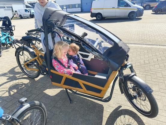 Two children sitting in a cargo bike, while an adult walks behind.