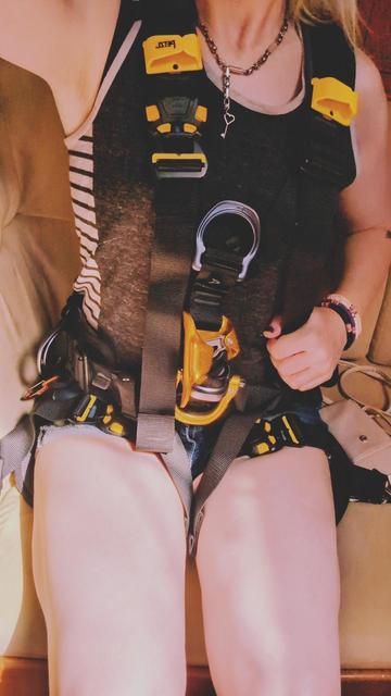 Alice, an athletic-looking blonde enby in a grey heathered tank top and denim shorty-shorts, is wearing a full harness that goes over their shoulders, around their waist, and between their thighs.