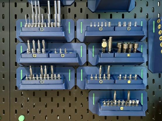 Seven 3D printed Gridfinity baseplates, a mix of three by one and four by one, are hanging from a SKÅDIS pegboard.  Each has one or two shallow bins containing metal screwdriver bits.  Each bin has a colored icon showing the kind of bit it holds (Phillips, Hex, Torx, etc.)