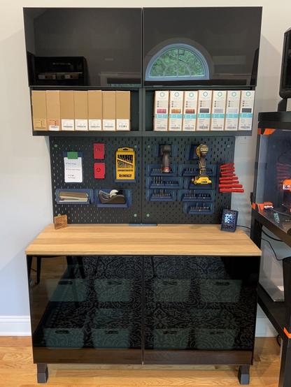 A tall cabinet is composed of two IKEA BESTÅ bottom and two top cabinets with glass doors.  Things are vaguely visible through the doors.  The bottom cabinets have an oak top.  Above that, there are two IKEA SKÅDIS pegboards.  The pegboards have a bunch of tools and things hanging from them.  The top cabinets have an open shelf below the doors, and each shelf holds 7 rolls of 3D printer filament in carboard boxes.  A 3D printer enclosure is to the right, mostly out of frame.