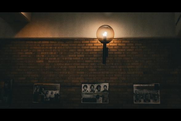 A dimly lit brick wall illuminated by a single globe wall sconce near the top center of the frame. Below the light, four vintage-style photo collages are mounted on the wall, showing portraits of people in black and white. The scene has a grainy, cinematic texture with warm tones from the light and dark shadows throughout.