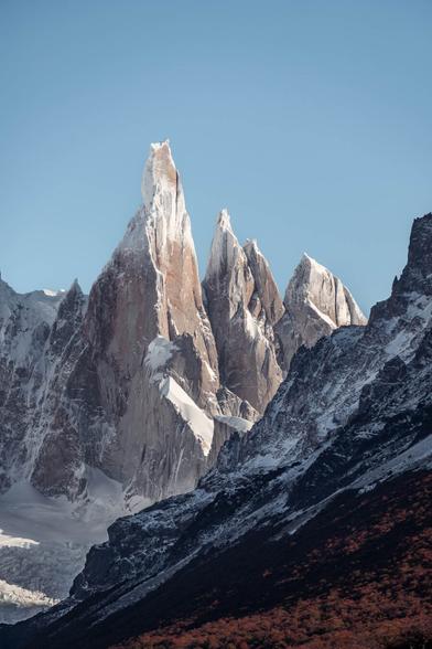 Dramatic view of Cerro Torre, a sharp spire-shaped mountain in Patagonia, rising above surrounding peaks. The scene is captured from a viewpoint near a glacial lagoon, with rugged terrain and partly cloudy skies emphasizing the remote, wild atmosphere.