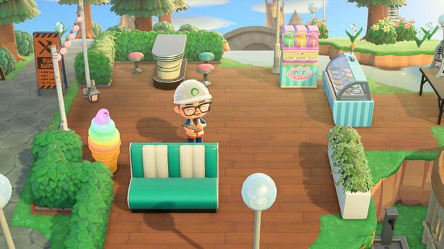 An Animal Crossing: New Horizons character in a hard hat stands on a wooden-paved plaza. New furniture for an ice cream parlor is visible, including a green and white diner bench, two pink stools, and what appears to be a soft-serve ice cream machine. In the background are more ice cream stalls, a large rainbow ice cream cone, and a yellow and black construction sign, indicating ongoing work.