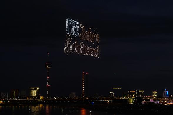 Der Schriftzug "175 Jahre Schlüssel", dargestellt mit 1000 Drohnen im Abendhimmel über dem Rhein in Düsseldorf. Im Hintergrund sieht man den Rheinturm und die Kniebrücke. Rechts im Bild ist ein kleiner Teil der Kirmes zu sehen.