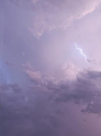Picture of a sky full of storm clouds. Above the biggest cloud there is one line shaped lightning. Sky is light colored and it has some light pink hues.