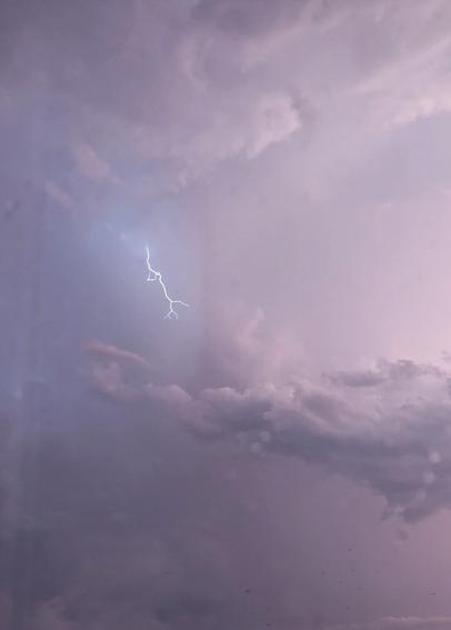 Picture of a sky full of storm clouds. On the left above the biggest cloud there is one line shaped lightning with couple small branches. Sky is light colored and it has some light pink hues.