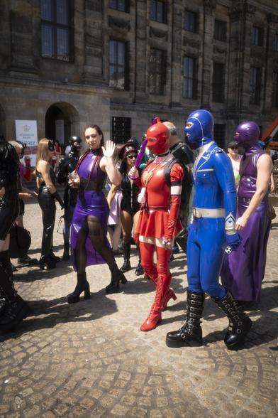A group of people dressed in colorful costumes featuring latex and leather materials stands on a cobblestone street in front of a historic building. Outfits include a red superhero-style suit with high boots, a blue suit with gloves, and a purple dress with high heels. Some wear masks while others have braided hair. Various accessories, such as a baton and utility belts, are visible. The street is busy with similarly dressed participants, suggesting a themed event or parade.