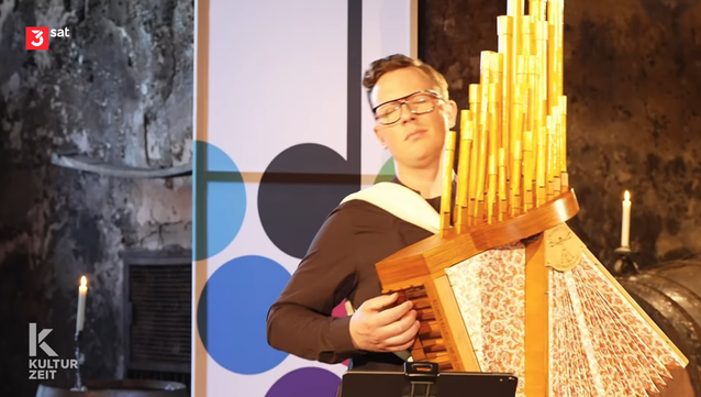 Screenshot aus dem Film
Der Akkordeonist spielt auf dem Ur-Akkordeon. Es ist ein Kasten aus Holz mit einem angebauten Balg, ähnlich dem bei modernen Akkordeons, aus dem oben ca. 25 verschieden lange Holzpfeifen ragen.