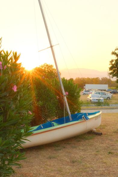 A small, narrow boat type vessel (don’t know much about these things, sorry) parked on a sandy beach between two bushes with a few pink flowers. Behind the bushes the sun is setting, creating a sun star effect in the camera. The vessel also has a tall metal rod I assume for the sails. It is so tall it goes out of frame. Behind all this there is a parking lot visible with a few cars and a mountain in the distance.