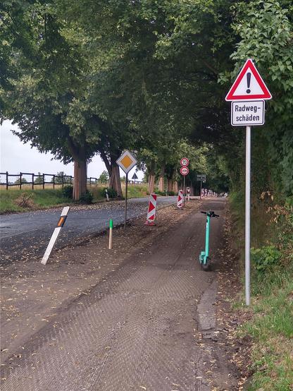 Baustelle an einer Landstraße. Es wird nicht nur die Autofahrbahn saniert, sondern auch der daneben verlaufende Radweg, an dem ein Schild vor "Radwegschäden" warnt.