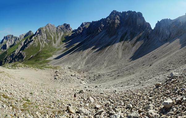 Im Vordergrund und in der Bildmitte ist ein steiniges Schuttfeld mit einer aufsteigenden Wanderin mit Rucksack zu sehen, das in eine Art Talkessel ausläuft und am Ende von teils im Schatten liegenden, steilen Bergflanken abgegrenzt wird. Weiter links geht es  immer grüner werdend weiter ins Tal hinunter.