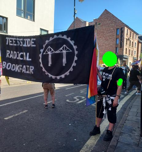 me with my face covered with a poison green blob carrying a black banner with 'Teesside Radical Bookfair' in silver letters with a cog circle with the tees suspension bridge in it as logo.
my black t-shirt says 'bob vylan is a sell-out'.